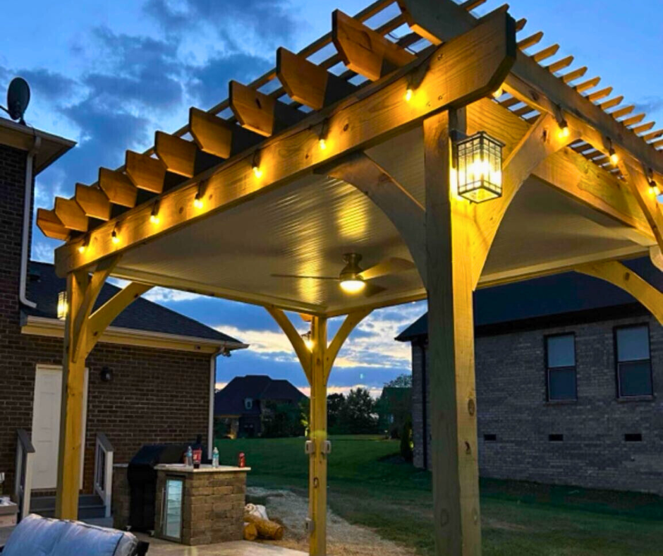 A wooden custom pergola with string lights and a fan, built by a pergola contractor.